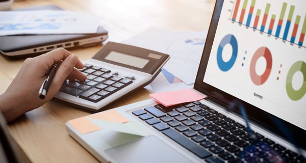 female hand using a calculator next to laptop covered in Post-It Notes with graphs and charts on the laptop screen all on a light brown wooden desk